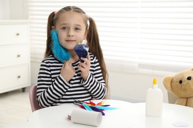 Photo of Little girl with paper bird and light blue feather indoors. Child handmade craft