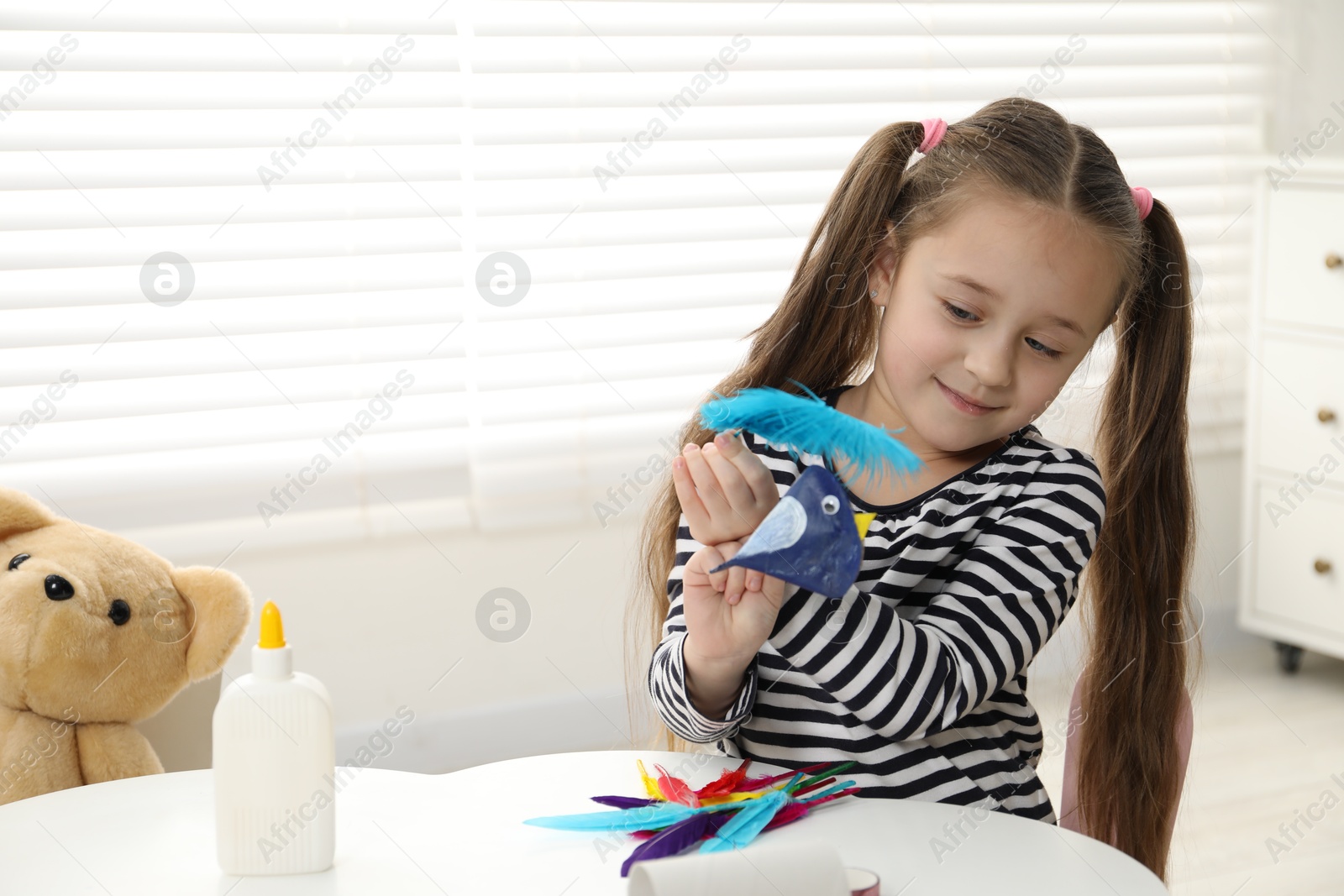 Photo of Little girl with paper bird and light blue feather indoors. Child handmade craft