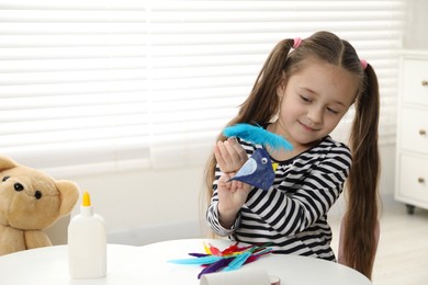 Photo of Little girl with paper bird and light blue feather indoors. Child handmade craft