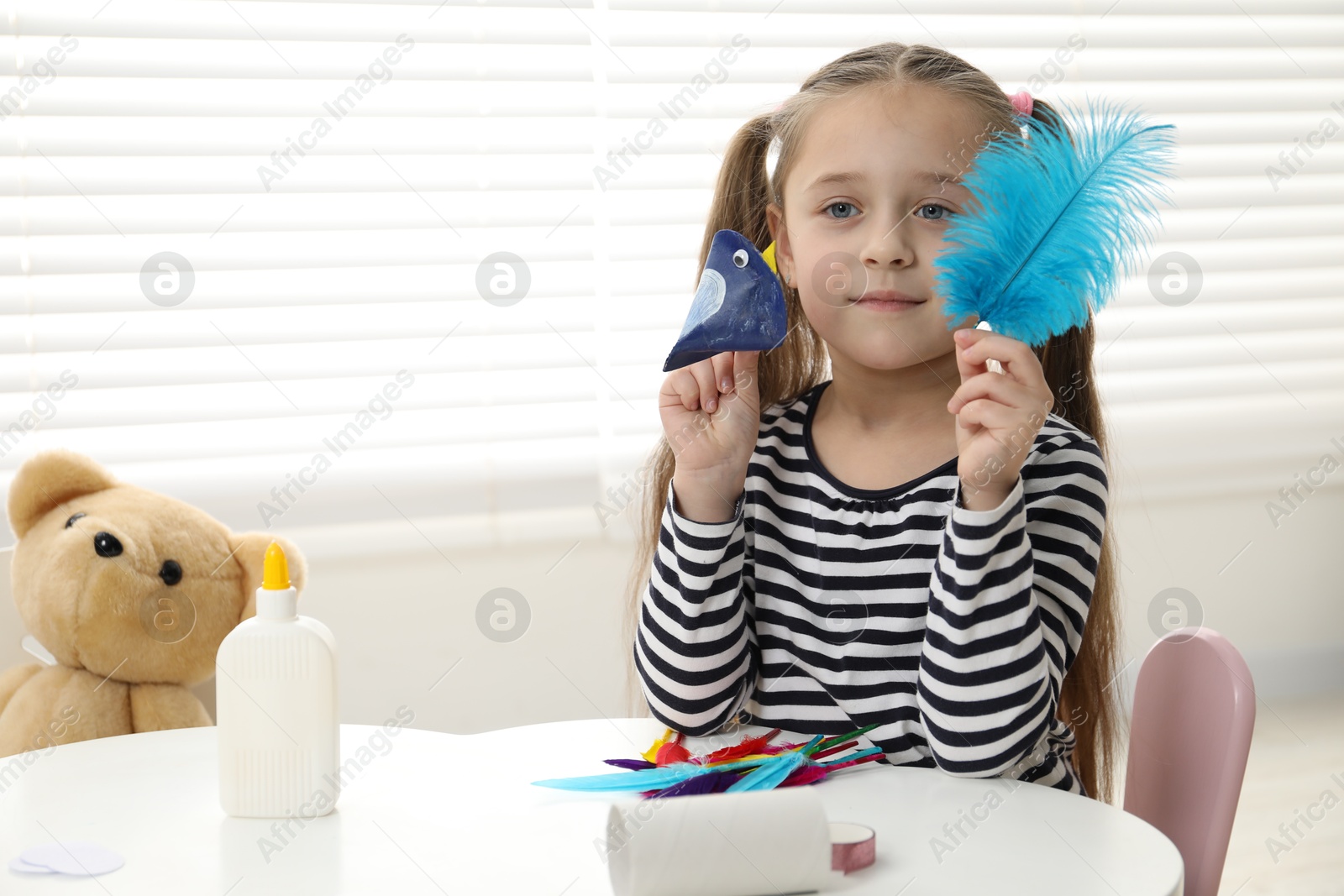 Photo of Little girl with paper bird and light blue feather indoors. Child handmade craft