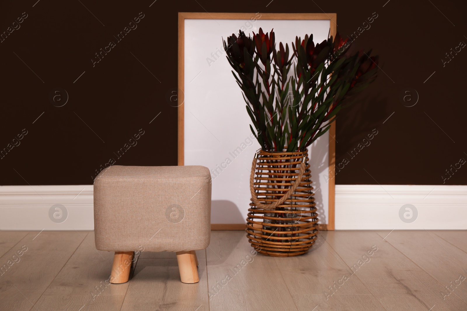 Photo of Stylish beige pouf, poster and decorative flowers near brown wall in room