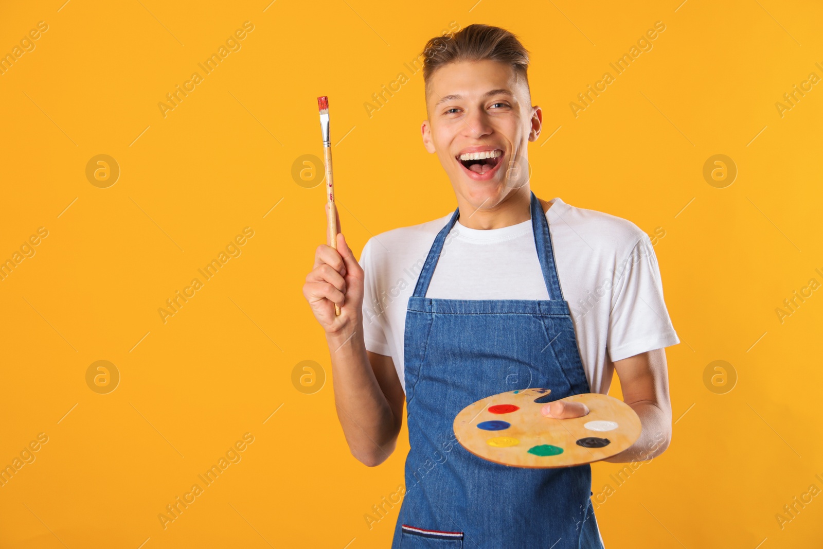 Photo of Happy man with wooden palette and paintbrush on orange background. Space for text