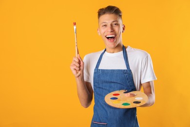 Photo of Happy man with wooden palette and paintbrush on orange background. Space for text