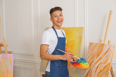 Smiling man with palette and paintbrush drawing picture indoors