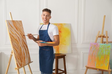 Smiling man with palette and paintbrush drawing picture indoors