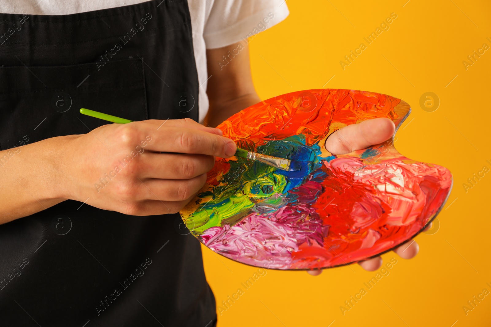 Photo of Man with paintbrush mixing paints on palette against orange background, closeup