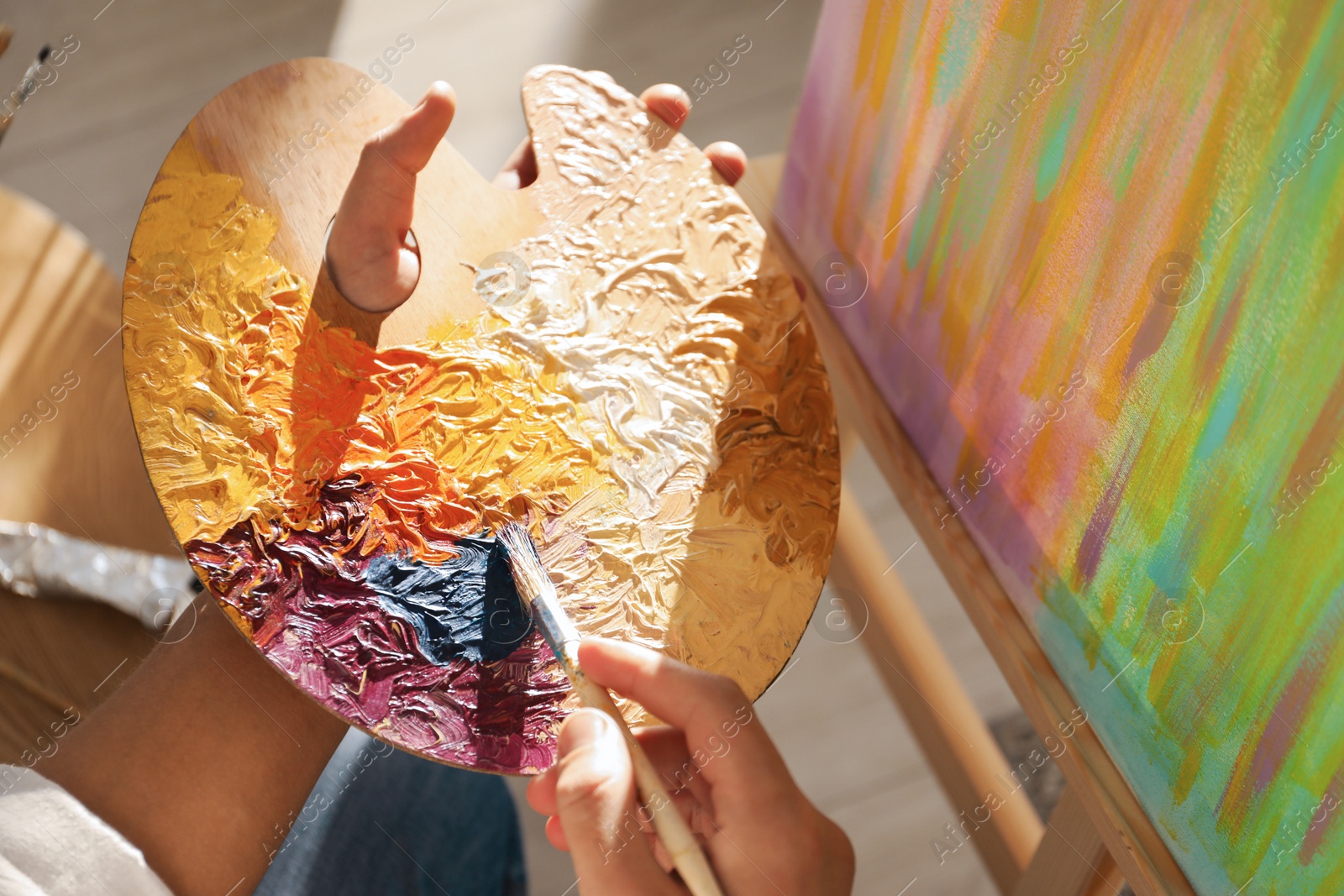 Photo of Man with paintbrush mixing paints on palette near picture indoors, closeup