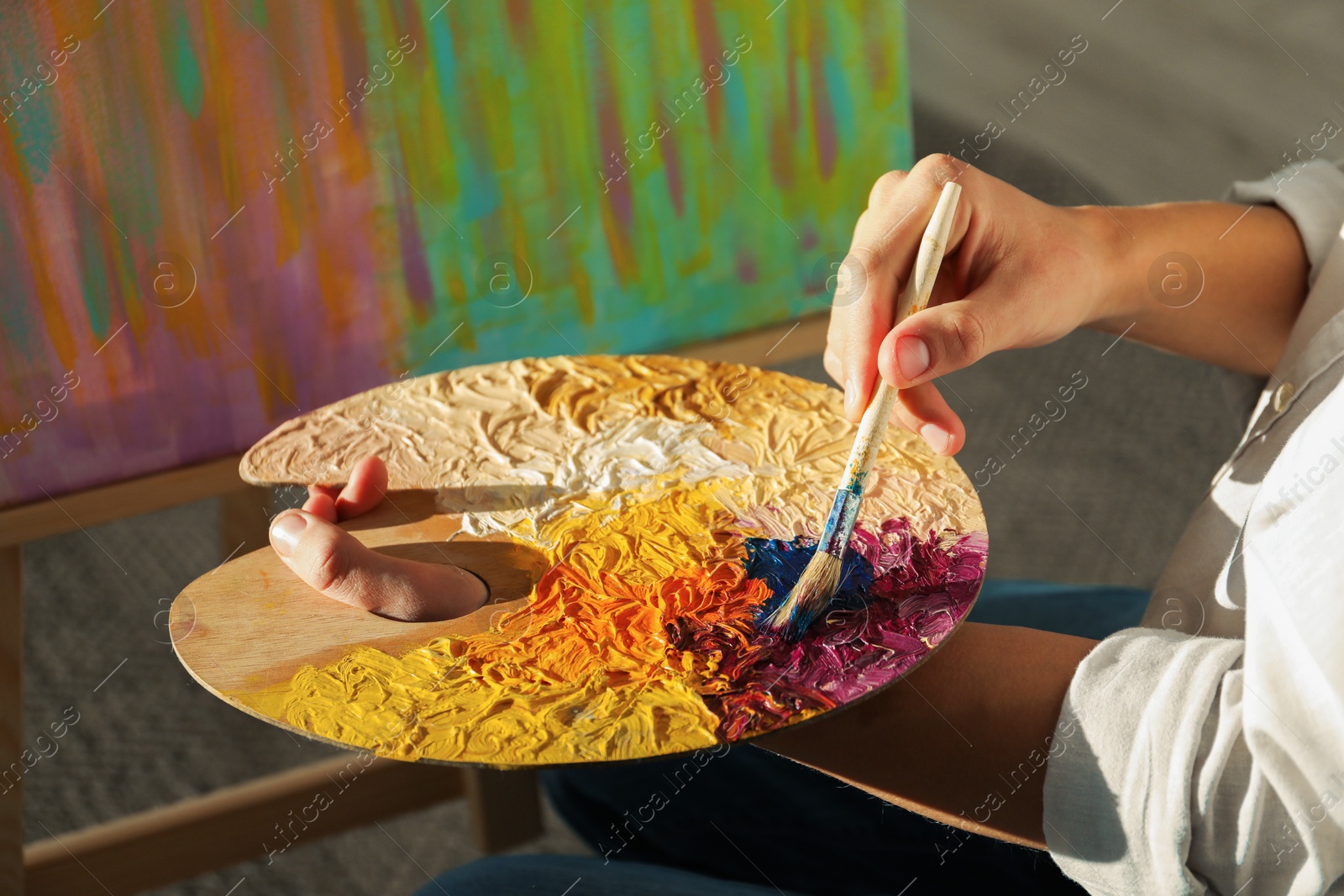 Photo of Man with paintbrush mixing paints on palette near picture indoors, closeup