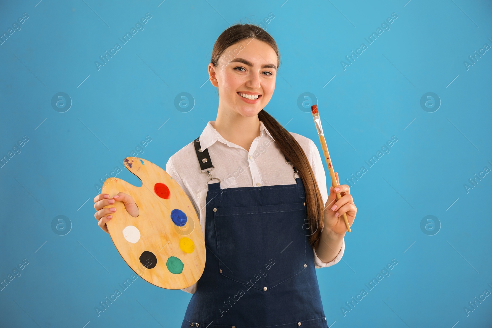 Photo of Smiling woman with wooden palette and paintbrush on light blue background