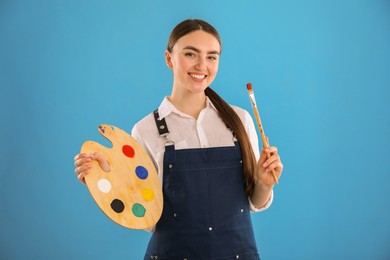 Photo of Smiling woman with wooden palette and paintbrush on light blue background