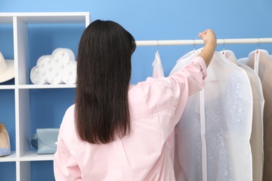 Photo of Woman taking garment cover with clothes from rack indoors, back view