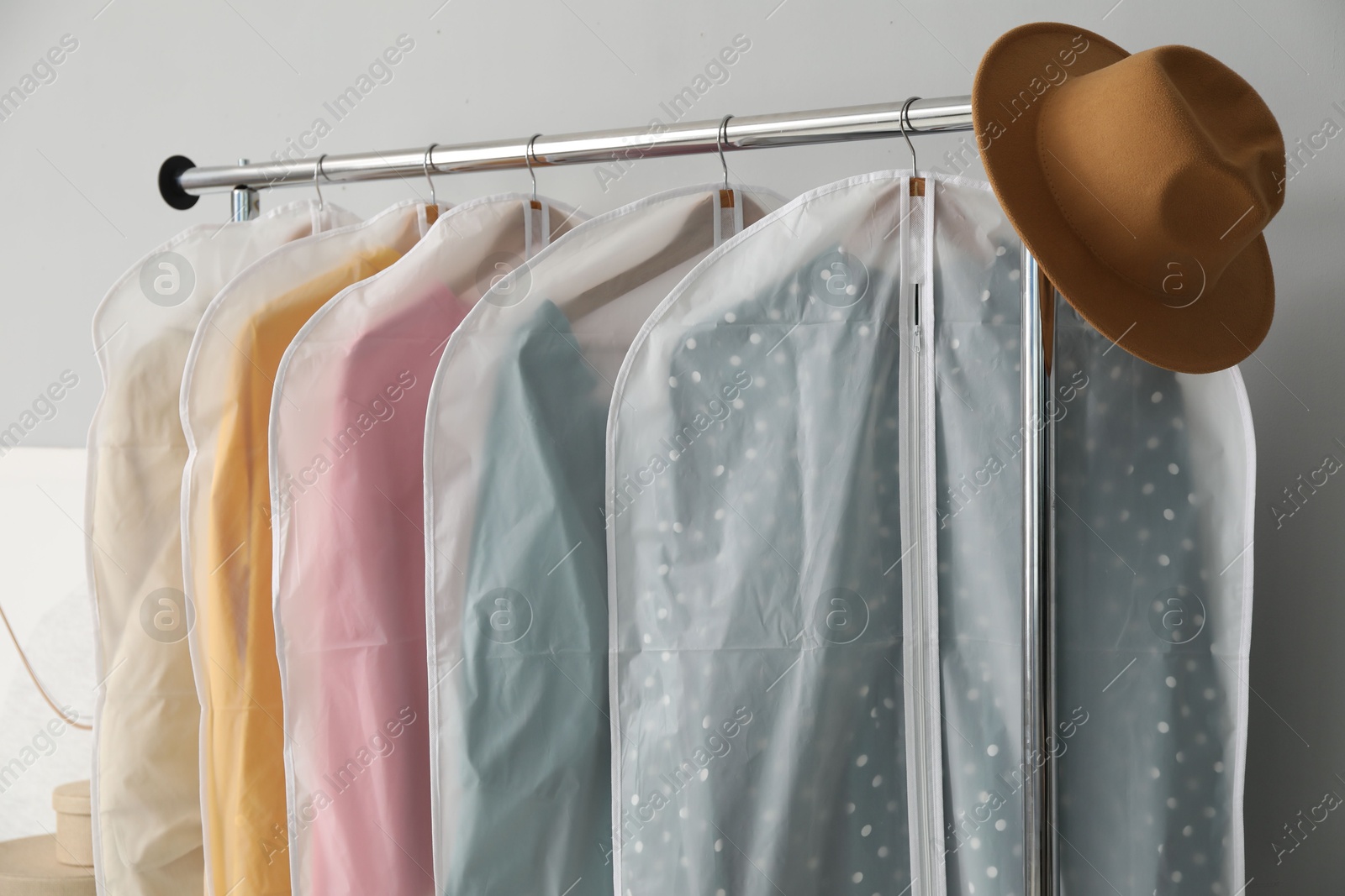 Photo of Garment covers with clothes and hat on rack near grey wall indoors