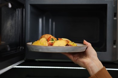 Photo of Woman putting plate with lunch into microwave indoors, closeup