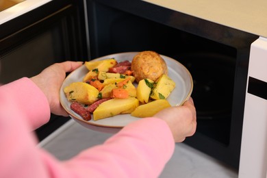 Photo of Woman putting plate with lunch into microwave indoors, closeup