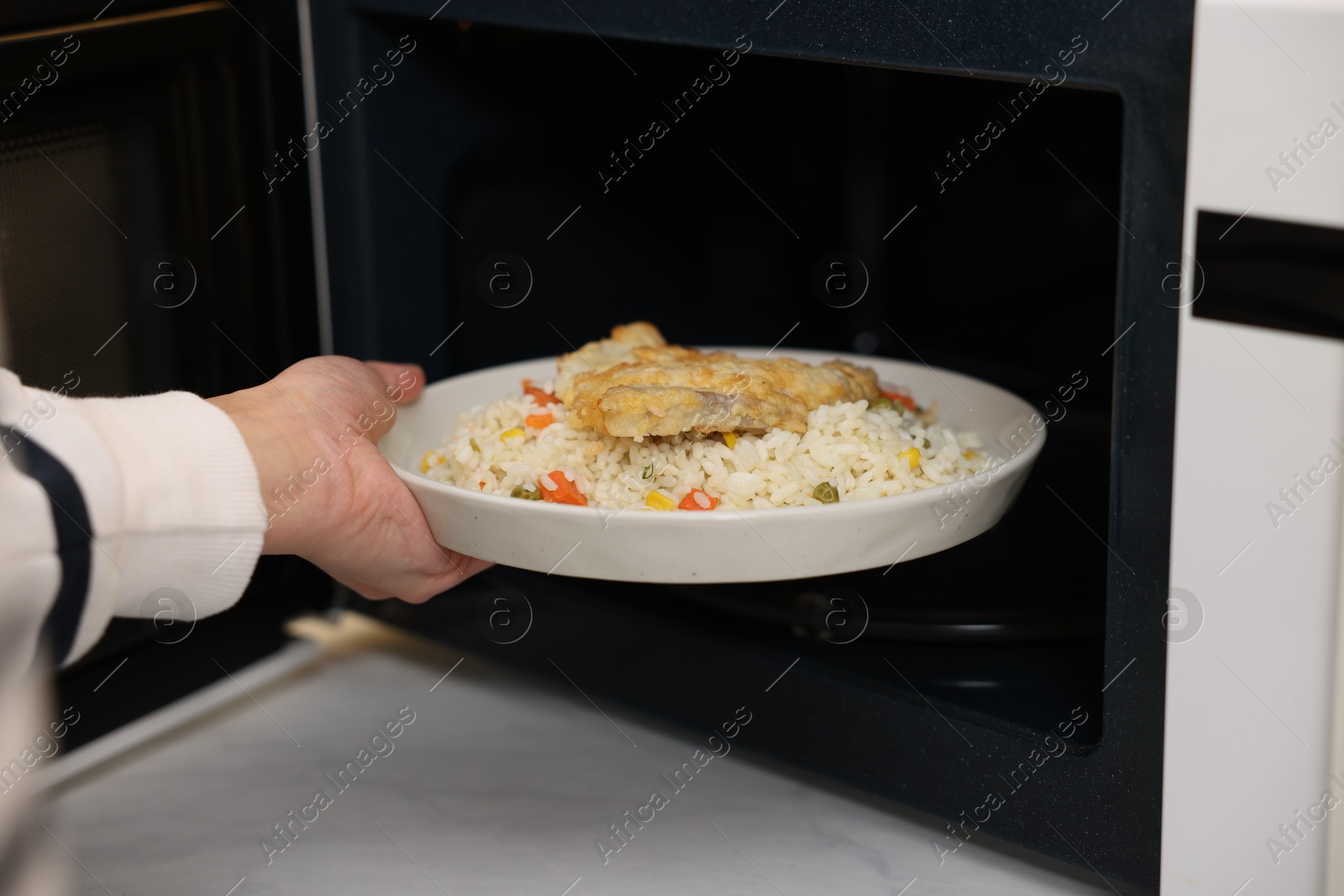 Photo of Woman putting plate with lunch into microwave indoors, closeup