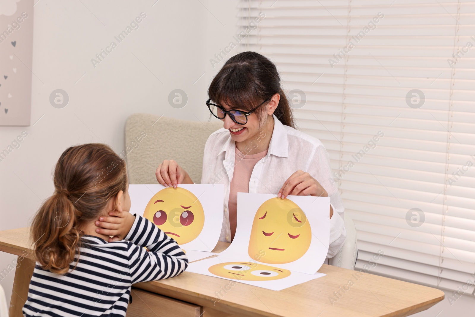 Photo of Autism therapy. Little girl choosing emoticon at table with smiling psychologist in mental health center