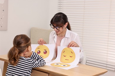 Photo of Autism therapy. Little girl choosing emoticon at table with smiling psychologist in mental health center