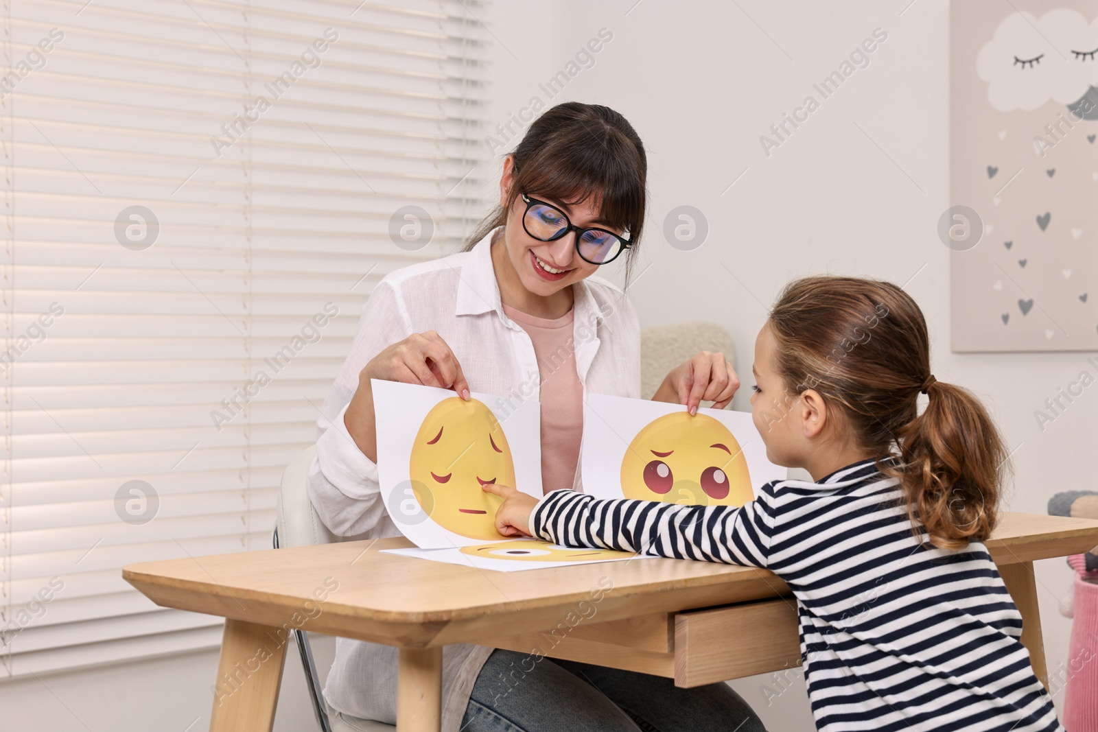 Photo of Autism therapy. Little girl choosing emoticon at table with smiling psychologist in mental health center