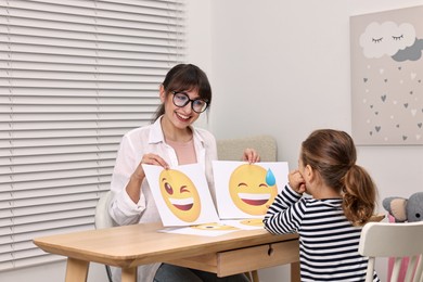 Photo of Autism therapy. Little girl choosing emoticon at table with smiling psychologist in mental health center