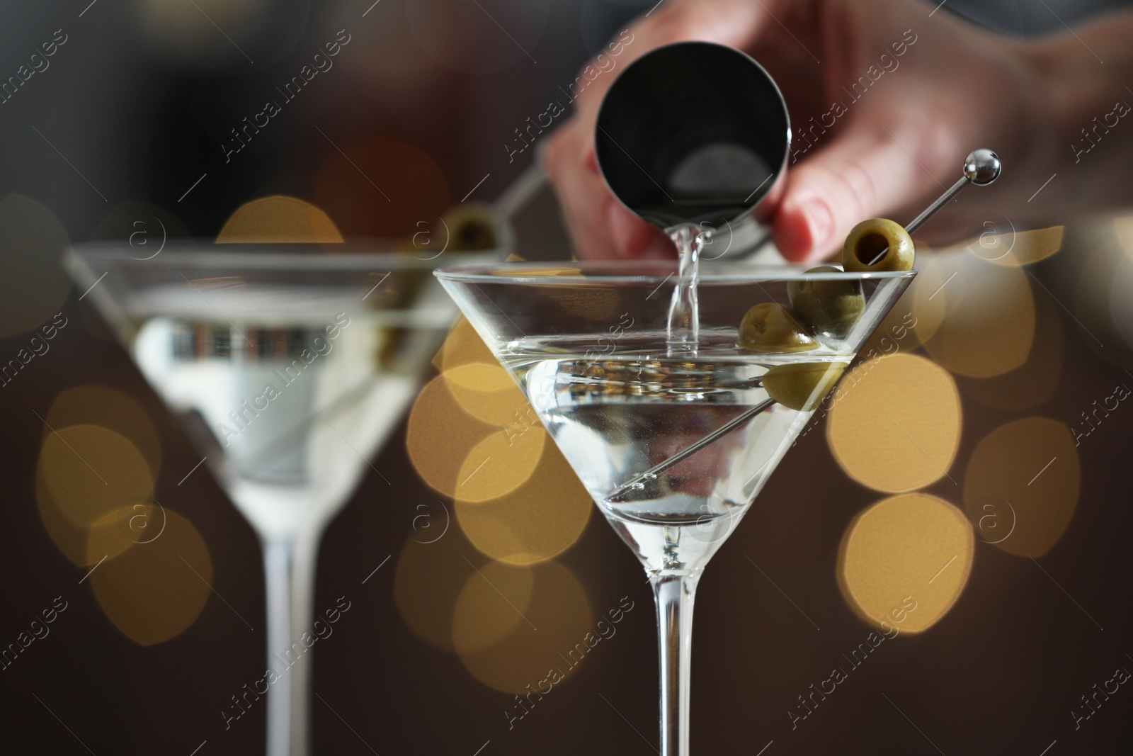 Photo of Woman making tasty martini cocktail with olives on blurred background, closeup. Bokeh effect