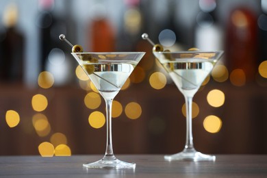 Photo of Martini cocktail with olives in glasses on wooden table against blurred background. Bokeh effect