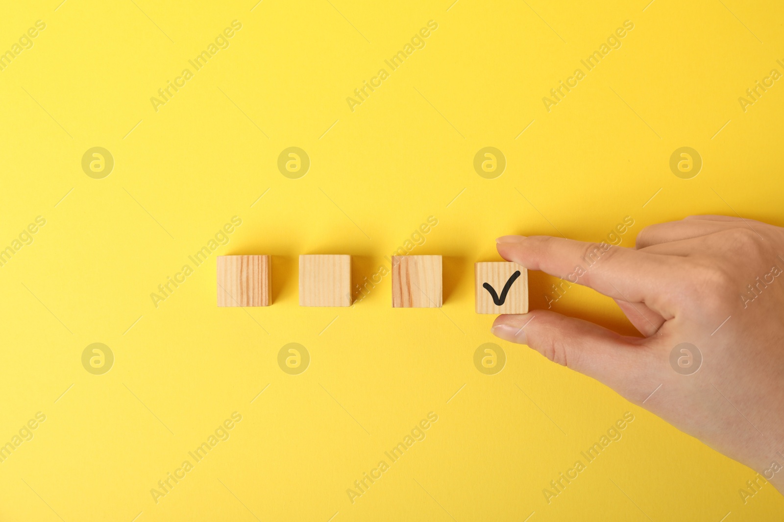 Photo of Woman taking wooden cube with check mark on yellow background, top view