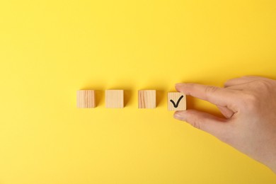 Photo of Woman taking wooden cube with check mark on yellow background, top view