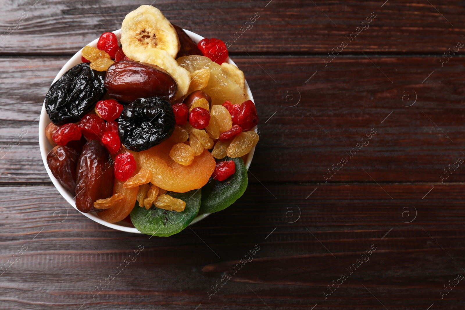 Photo of Mix of different dried fruits in bowl on wooden table, top view. Space for text