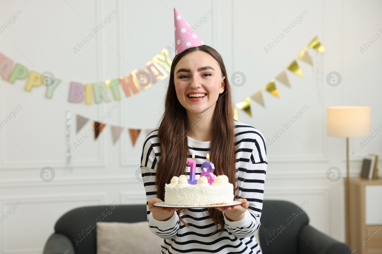 Photo of Coming of age party - 18th birthday. Happy young woman in hat holding tasty cake with number shaped candles at home