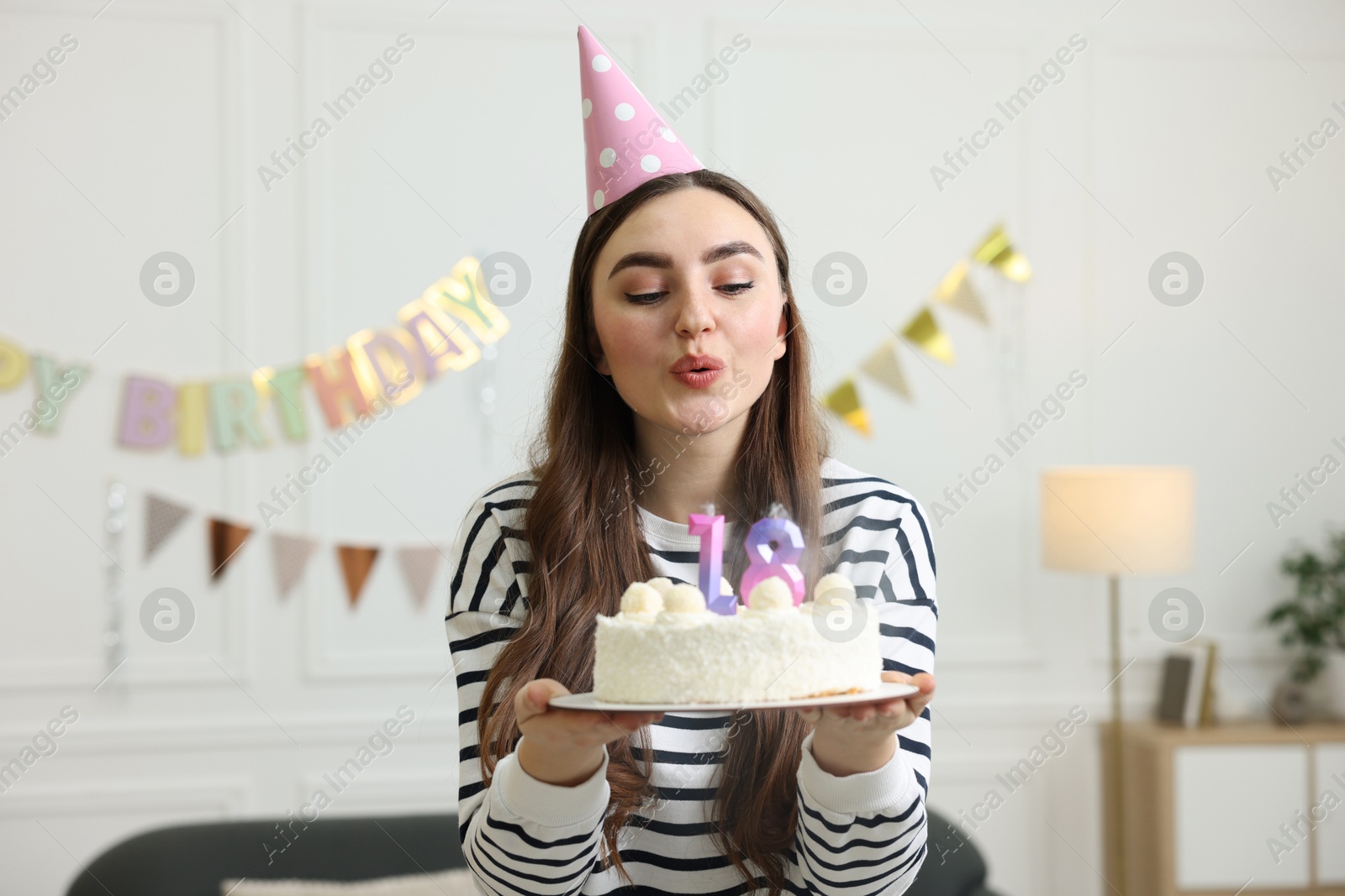 Photo of Coming of age party - 18th birthday. Young woman in hat holding tasty cake and blowing out number shaped candles at home