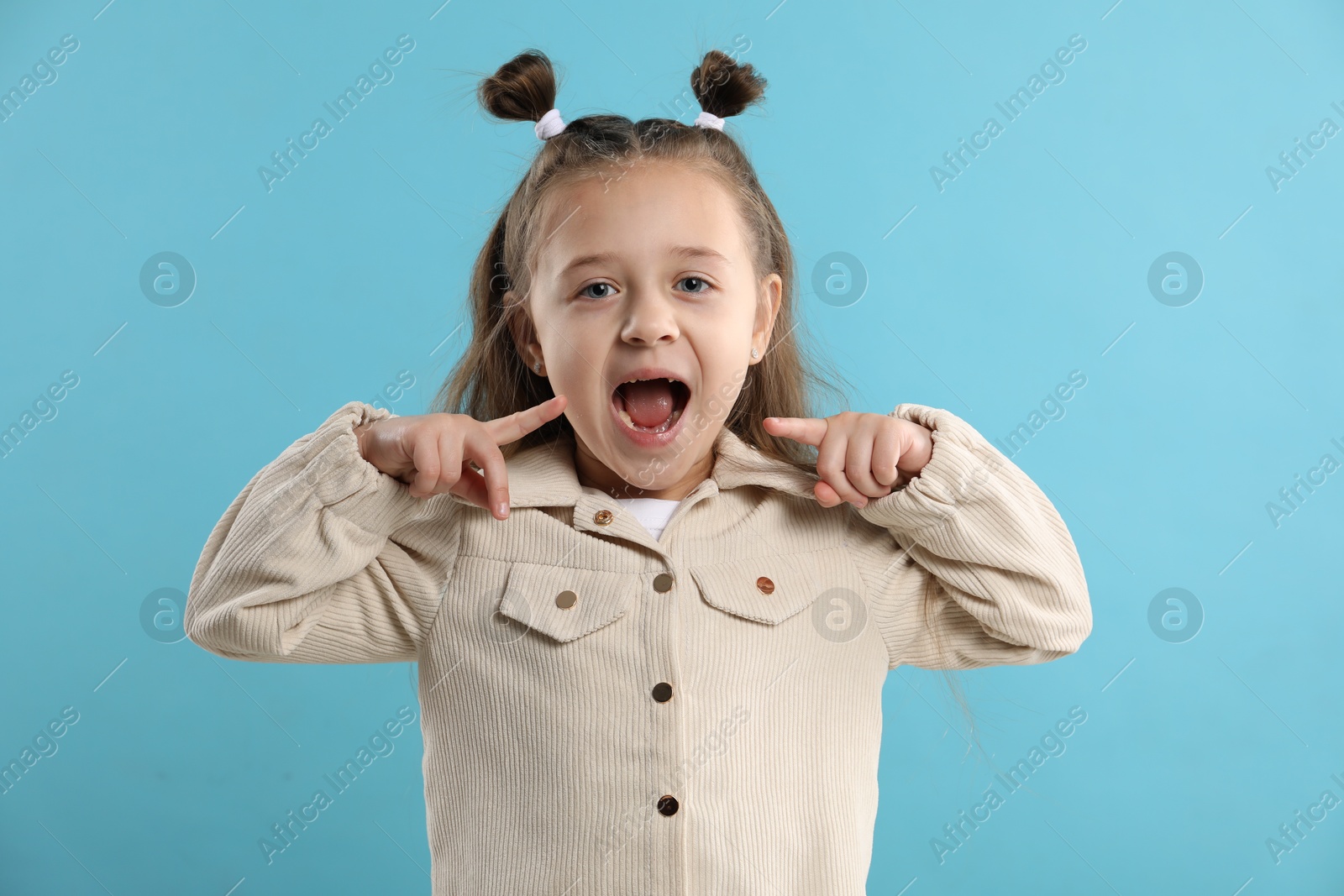 Photo of Cute little girl pointing at her missing tooth on light blue background. Waiting for tooth fairy