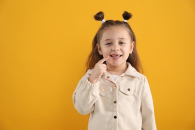 Photo of Cute little girl pointing at her missing tooth on orange background, space for text. Waiting for tooth fairy