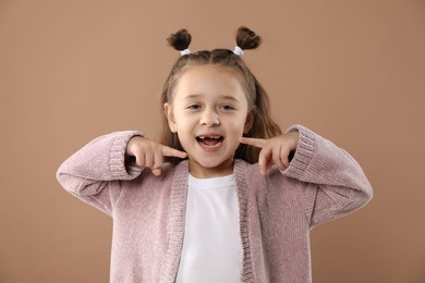 Photo of Cute little girl pointing at her missing tooth on light brown background. Waiting for tooth fairy