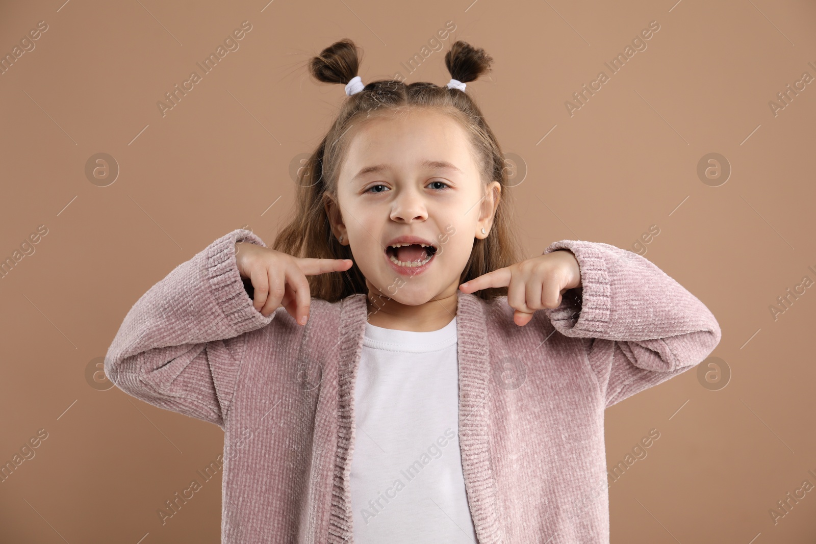 Photo of Cute little girl pointing at her missing tooth on light brown background. Waiting for tooth fairy