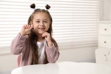 Photo of Cute little girl with missing tooth at white table indoors, space for text. Waiting for tooth fairy