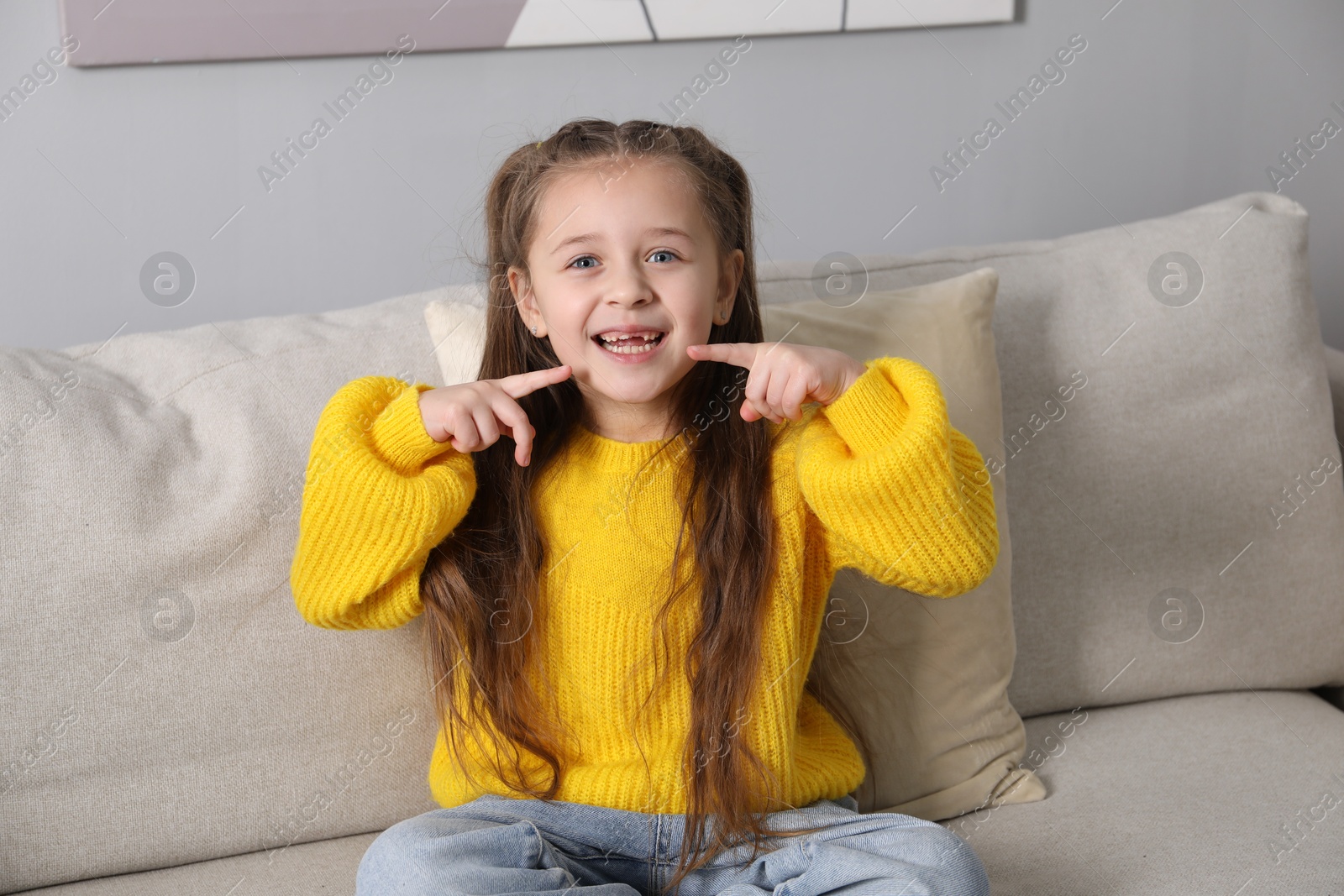 Photo of Cute little girl pointing at her missing tooth on sofa indoors. Waiting for tooth fairy