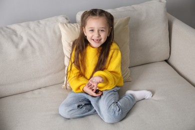 Photo of Cute little girl with missing tooth on sofa indoors. Waiting for tooth fairy