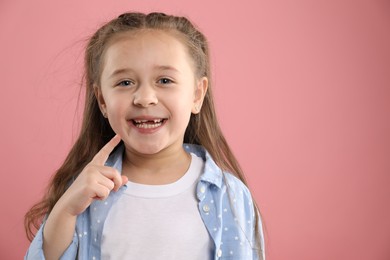 Photo of Cute little girl pointing at her missing tooth on pink background, space for text. Waiting for tooth fairy