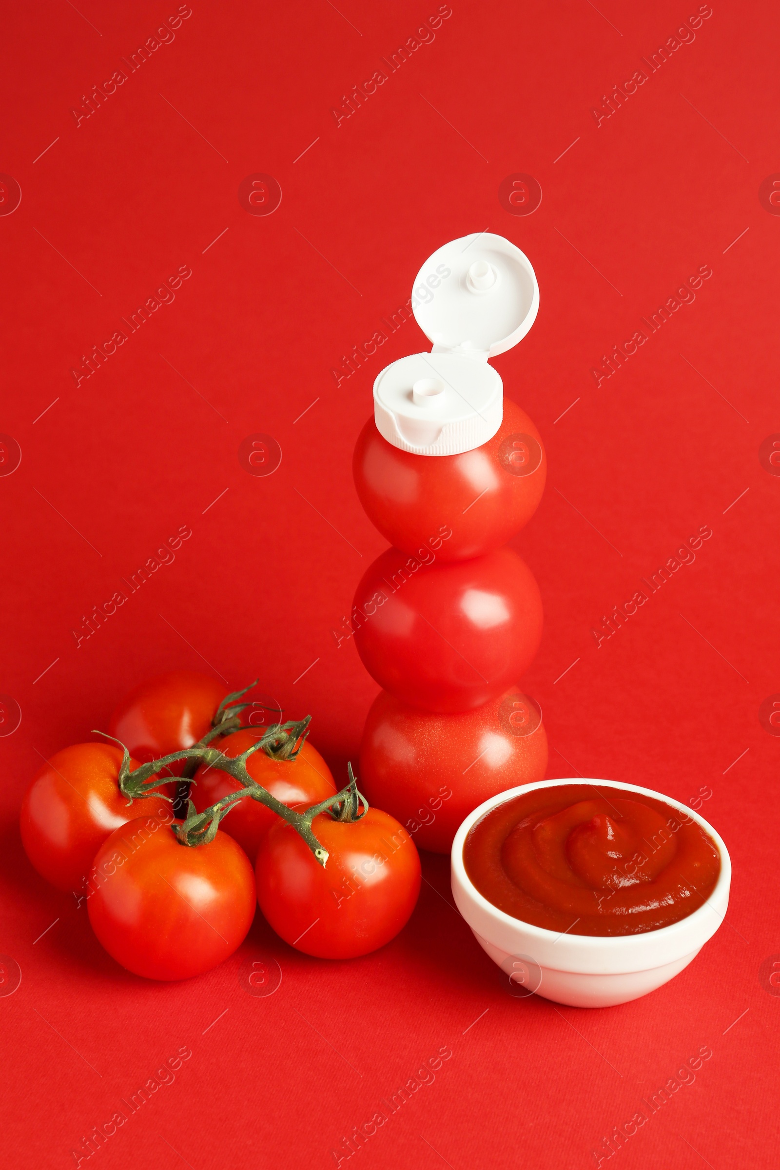 Photo of Stack of fresh tomatoes with plastic cap as bottle and ketchup in bowl on red background