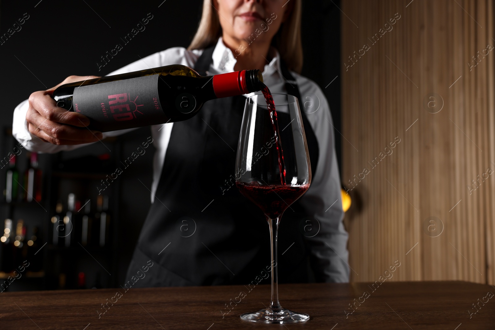 Photo of Professional sommelier pouring red wine into glass at wooden table indoors, closeup