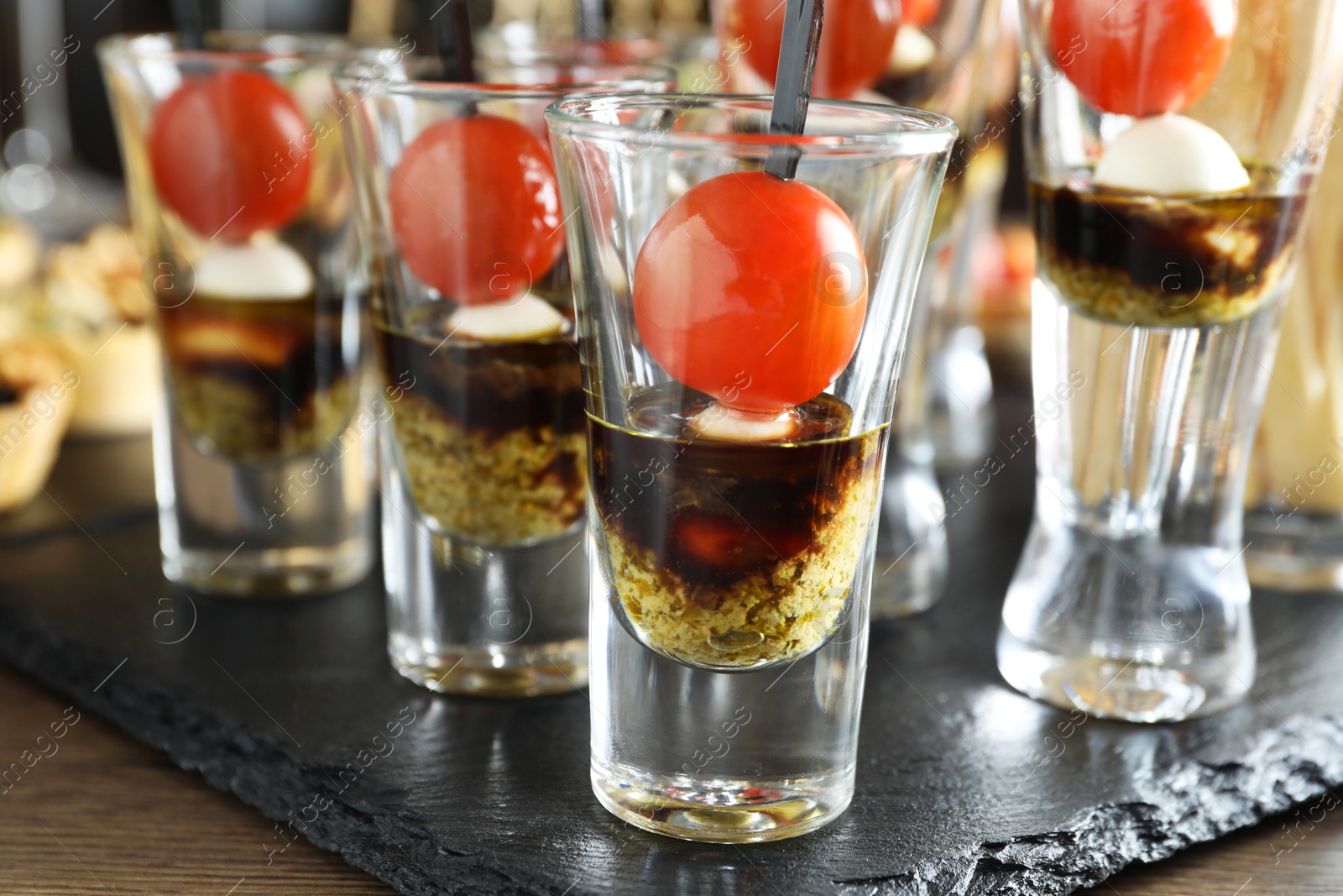 Photo of Tasty canapes with cherry tomatoes, mozzarella, balsamic vinegar and pesto sauce in shot glasses on wooden table, closeup