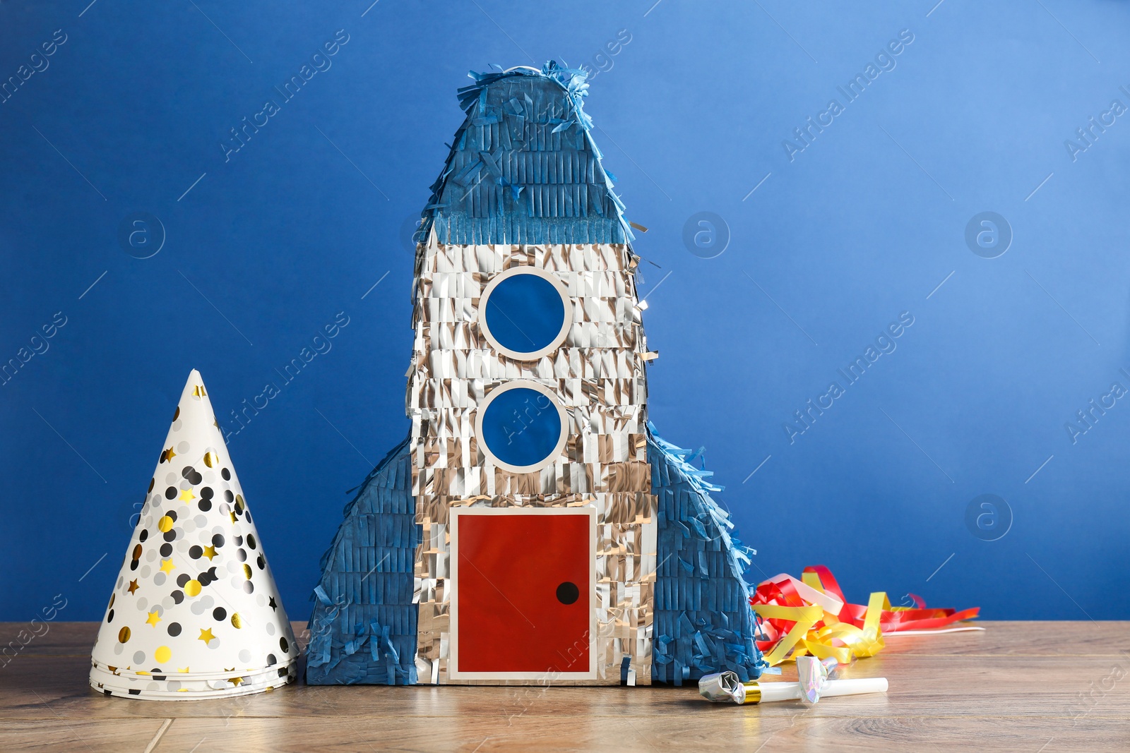 Photo of Beautiful pinata in shape of rocket, party cones and blowers on wooden table against blue background