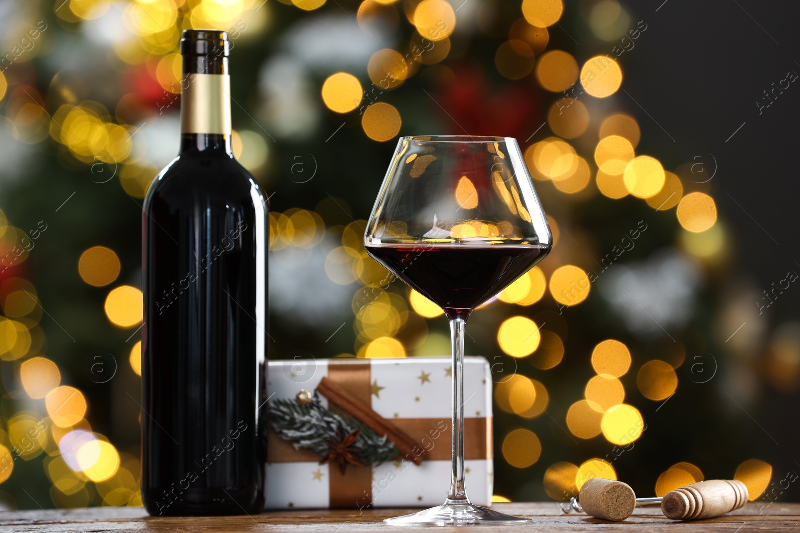 Photo of Red wine in glass, bottle, corkscrews and Christmas gift on wooden table against blurred lights