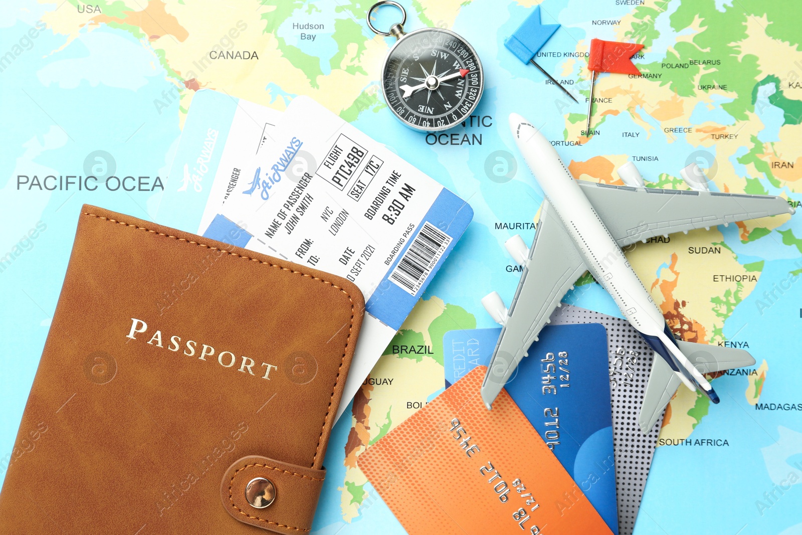 Photo of Passport in brown cover, flight tickets, plane model, compass, credit cards and small flags on world map, flat lay