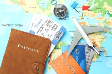 Photo of Passport in brown cover, flight tickets, plane model, compass, credit cards and small flags on world map, flat lay