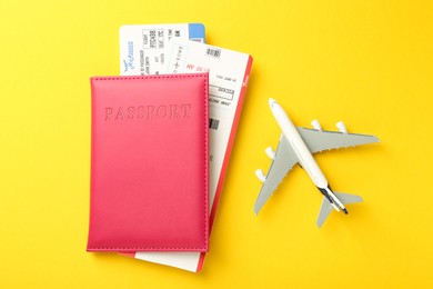 Photo of Passport in pink cover, plane model and flight tickets on yellow background, top view