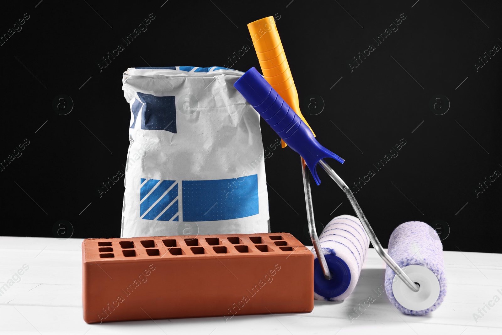 Photo of Brick, bag of cement and paint rollers on white wooden table