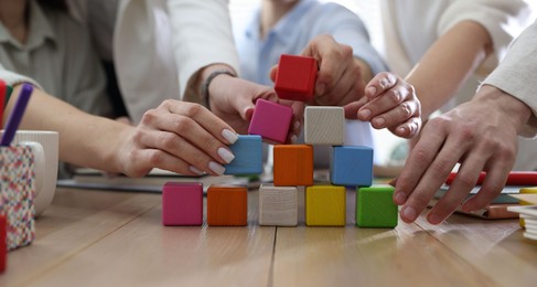 Unity concept. People building pyramid of colorful cubes at wooden table indoors, closeup