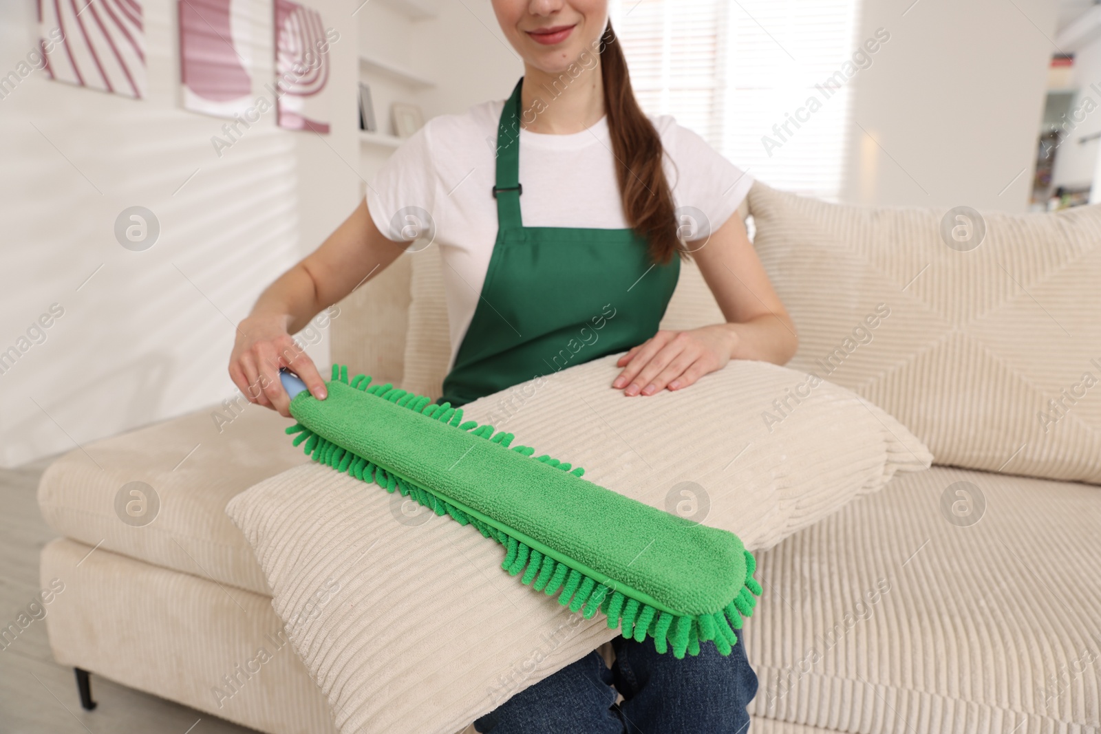 Photo of Janitor cleaning sofa cushion with duster at home, closeup