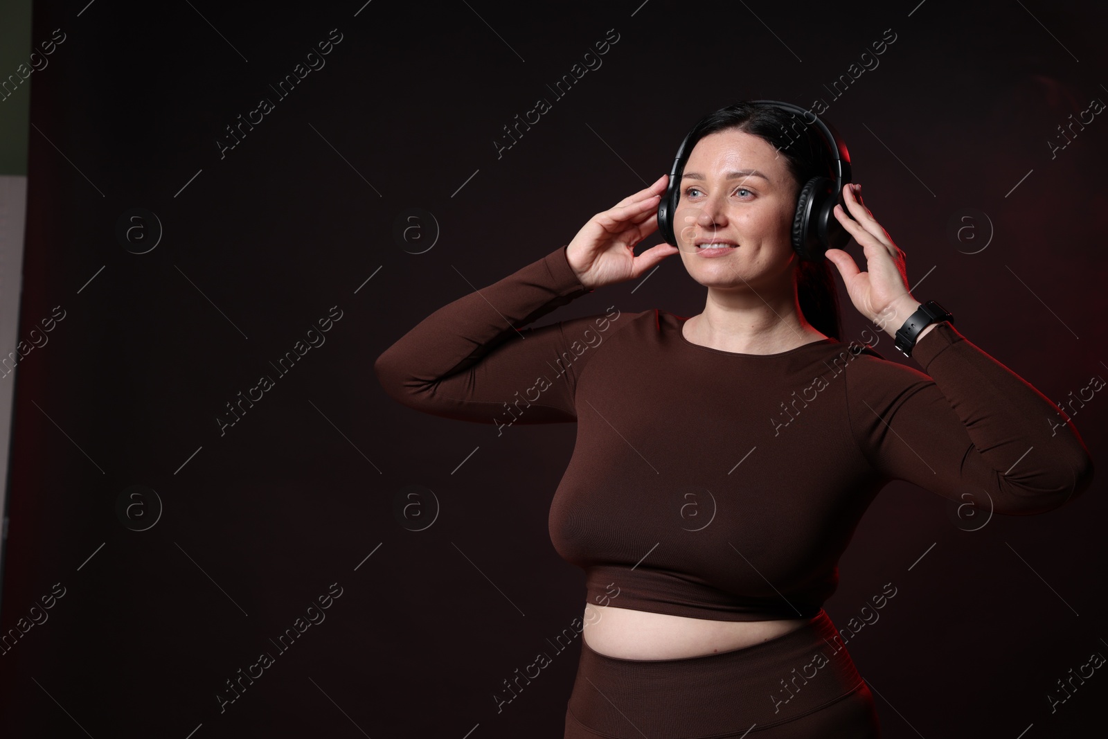 Photo of Plus size woman in gym clothes and headphones on black background with smoke, space for text